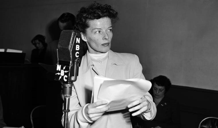 A black and white photo of Katharine Hepburn holding a sheaf of papers, preparing to speak into a NBC microphone.